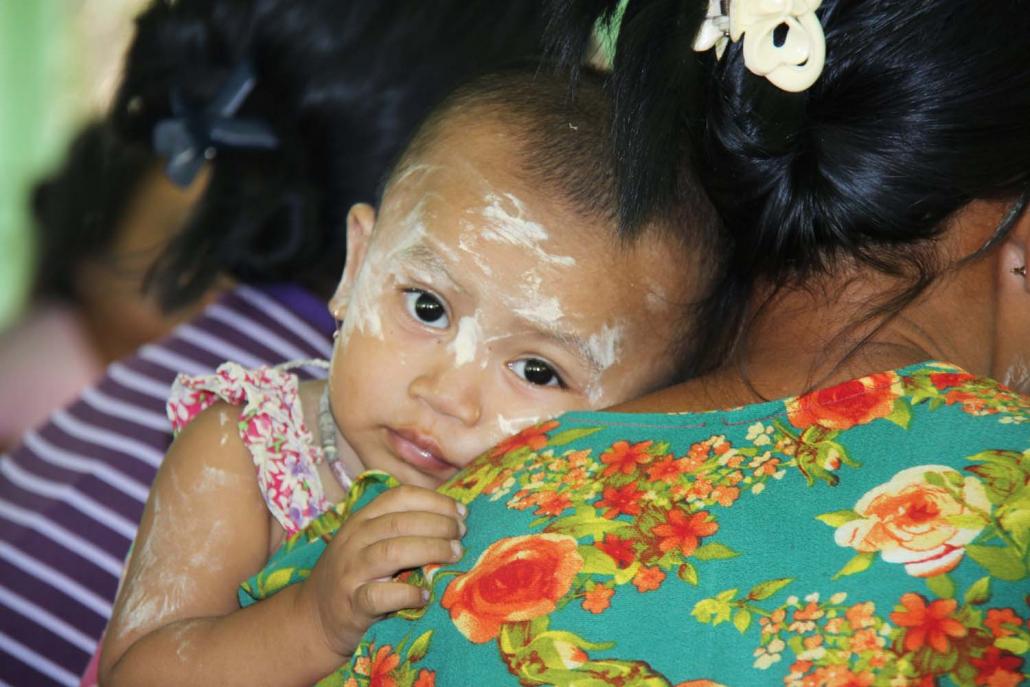 A child looks over her mother’s shoulder in Myint Gar Thit village, Pauktaw Township, during a meeting between Tat Lan MCCT recipients and the Department of Social Welfare. (Kaung Htet | Tat Lan)