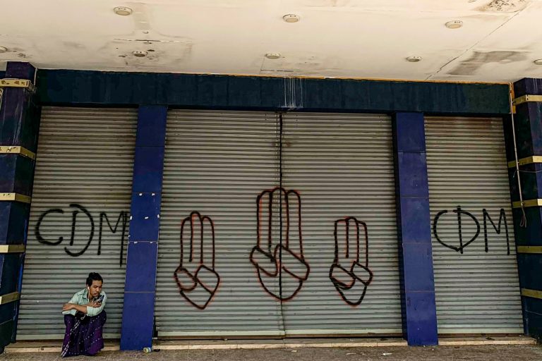 A man in Yangon on April 7 sits in front of shutters with the graffitied initials of the Civil disobedience Movement and the three-finger salute made by anti-coup protesters. (AFP)